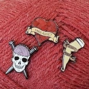 May include: Three enamel pins featuring a skull with knitting needles, a heart with the text "Yarn Addict", and a yarn ball with knitting needles. The pins are on a red ball of yarn.