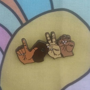 May include: A gold-toned enamel pin depicting three hands forming the sign language symbol for "love". The hands are different skin tones: brown, light brown, and tan.