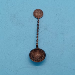 May include: A silver spoon with a twisted handle and a round bowl. The bowl and the top of the handle are decorated with a coin featuring a cross.