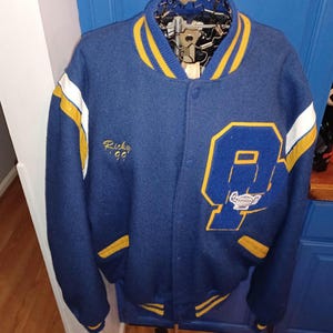 May include: A blue varsity jacket with yellow and white accents. The jacket features a large letter "A" on the front, along with the name "Ricky" and the year "99" embroidered in gold. The collar and cuffs have yellow stripes.