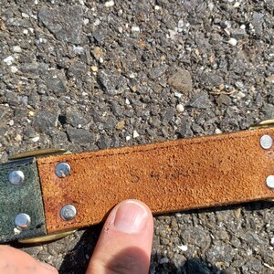 以下が含まれることがあります： 茶色の革ベルトのアップで、緑色の革の部分があります。ベルトには真鍮製のバックルと銀色のリベットが付いています。革は使い込まれていて、ビンテージな雰囲気です。ベルトには「S 4185」という文字が刻印されています。