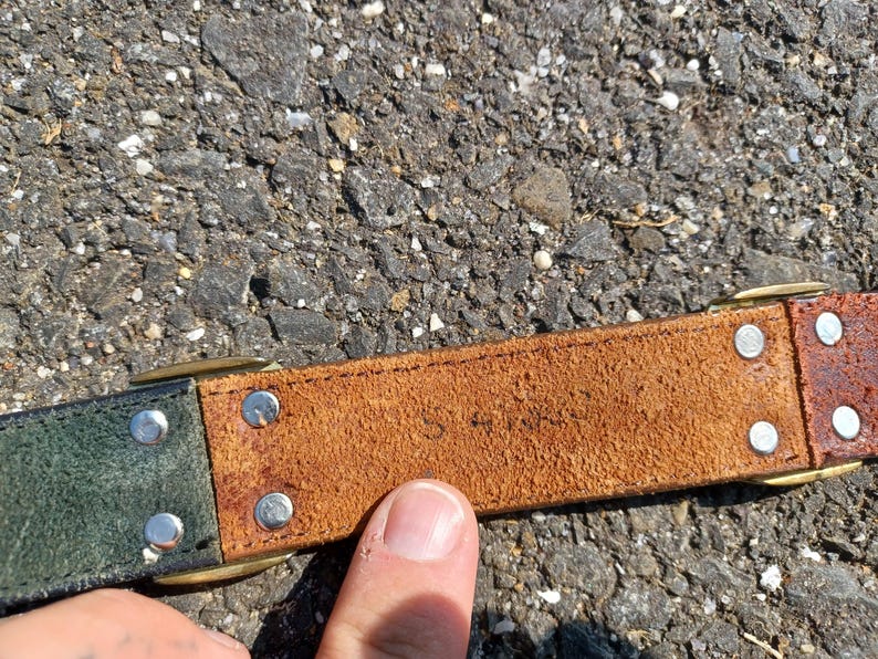 以下が含まれることがあります： 茶色の革ベルトのアップで、緑色の革の部分があります。ベルトには真鍮製のバックルと銀色のリベットが付いています。革は使い込まれていて、ビンテージな雰囲気です。ベルトには「S 4185」という文字が刻印されています。