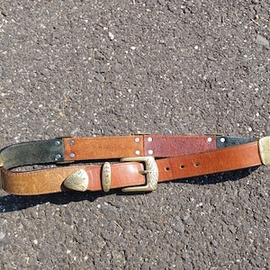 以下が含まれることがあります： 真鍮のバックルと装飾的なデザインが施された茶色の革ベルト。ベルトは3種類の茶色の革で作られており、濃い緑色の革のストリップが付いています。