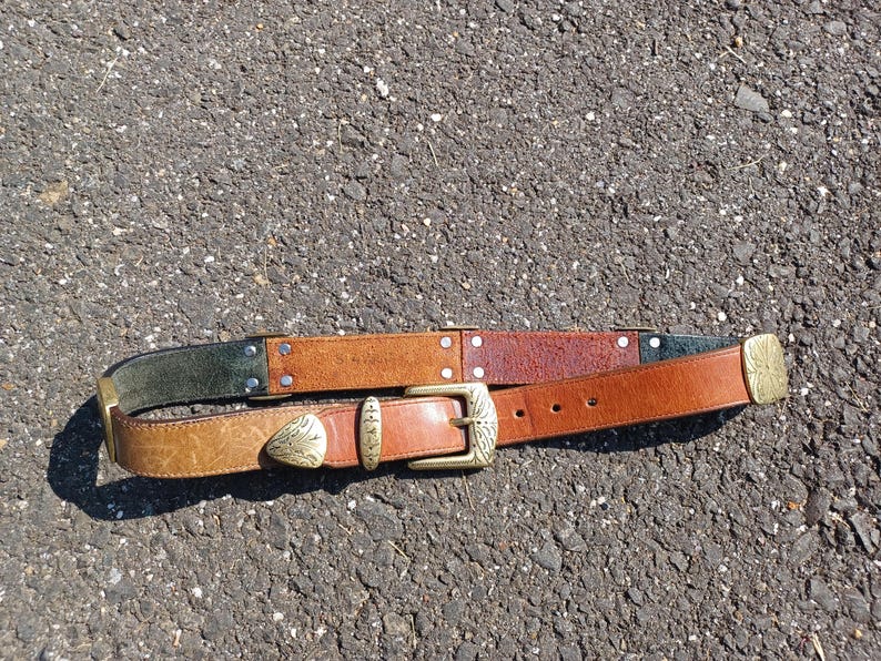 以下が含まれることがあります： 真鍮のバックルと装飾的なデザインが施された茶色の革ベルト。ベルトは3種類の茶色の革で作られており、濃い緑色の革のストリップが付いています。