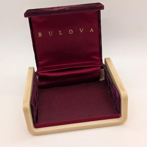 May include: Open Bulova watch box with a burgundy velvet interior and a beige exterior. The brand name "BULOVA" is displayed in gold lettering on the inside lid. The box is designed to hold a watch.