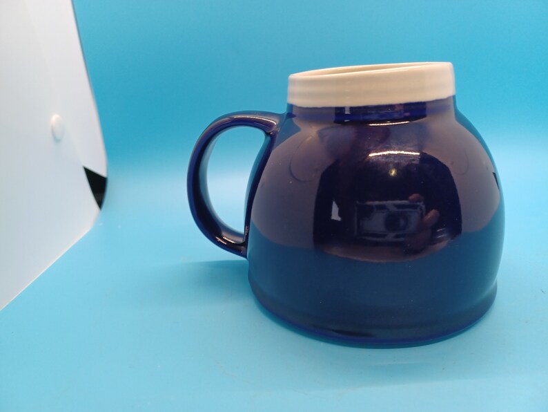 Pu&ograve; includere: Una tazza in ceramica blu scuro con un bordo bianco. La tazza ha una forma arrotondata e un manico.