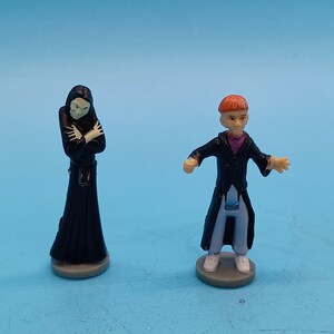 May include: Two small plastic figurines, one in a black robe with a white face and the other in a black coat with a red hat and a white face.