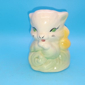 May include: A white ceramic planter shaped like a cat with green eyes and a yellow flower on its head. The cat is sitting with its paws crossed and has a green and white swirl pattern on its body.
