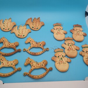 May include: A collection of 12 vintage Christmas ornaments featuring a variety of designs, including rocking horses, doves, and snowmen. The ornaments are made of a light brown material and are decorated with green, red, and white paint.