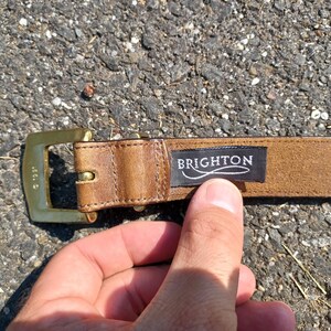 以下が含まれることがあります： ゴールドのバックルと「BRIGHTON」と書かれた黒い革のラベルが付いた茶色の革ベルトです。