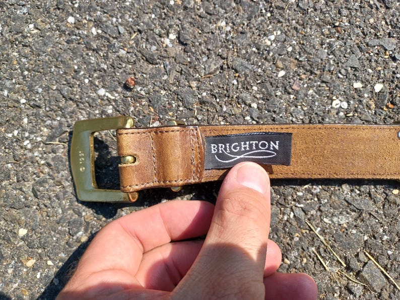 以下が含まれることがあります： ゴールドのバックルと「BRIGHTON」と書かれた黒い革のラベルが付いた茶色の革ベルトです。
