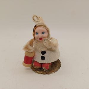 May include: A vintage snowman figurine with a cream-colored coat, a matching hat, and red boots. The snowman has a painted face with blue eyes and a red mouth. It holds a red and tan lantern. The base is gold-colored.