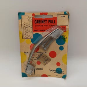 May include: A silver cabinet pull with screws, packaged on a colorful card with the text "CABINET PULL COMPLETE WITH SCREWS HEAVY PLATE ON CABINET" and "UNCONDITIONALLY GUARANTEED".