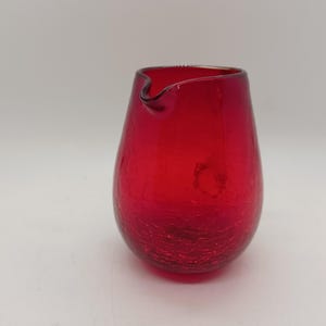 May include: A small, red glass pitcher with a textured surface. The pitcher has a rounded body and a slightly flared lip.