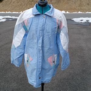 Puede incluir: Una chaqueta azul claro con un diseño de patchwork blanco y rosa. La chaqueta tiene una cremallera y un cuello.