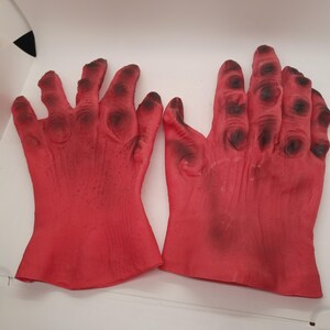 May include: A pair of red rubber monster hands with black detailing. The hands are outstretched with fingers spread apart.