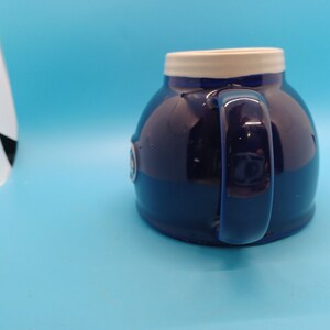 Pu&ograve; includere: Una tazza in ceramica blu con un bordo bianco e un logo bianco sul lato. La tazza ha una forma arrotondata e un manico.