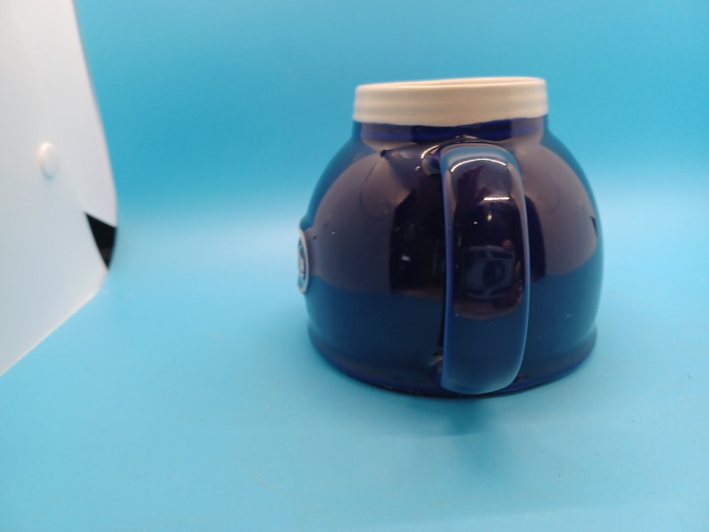 Pu&ograve; includere: Una tazza in ceramica blu con un bordo bianco e un logo bianco sul lato. La tazza ha una forma arrotondata e un manico.