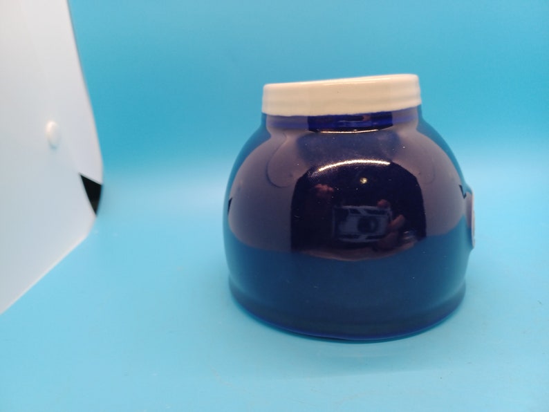 Pu&ograve; includere: Un vaso di ceramica blu scuro con un coperchio bianco. Il vaso ha una finitura liscia e lucida.
