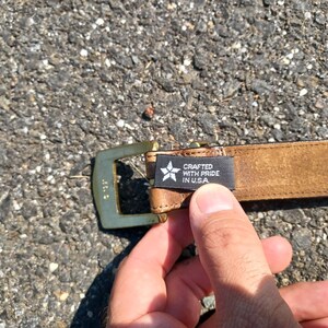 以下が含まれることがあります： ゴールドのバックルと「Crafted with Pride in USA」と書かれた黒いラベルが付いた茶色の革ベルトです。