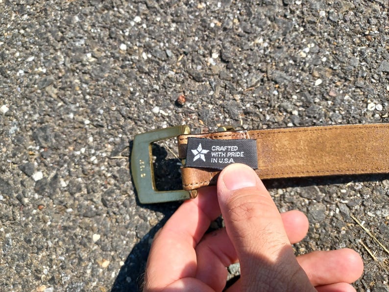 以下が含まれることがあります： ゴールドのバックルと「Crafted with Pride in USA」と書かれた黒いラベルが付いた茶色の革ベルトです。