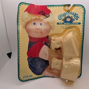 May include: A Cabbage Patch Kids doll shoes and socks set in original packaging. The cardboard backing features a blonde doll in a red shirt and blue overalls. The plastic blister pack contains a pair of cream-colored shoes with laces and socks.