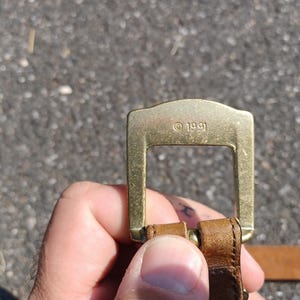 以下が含まれることがあります： 長方形で滑らかな表面を持つ、金色の金属製のベルトバックル。バックルには「&copy; 1991」という小さな刻印があります。