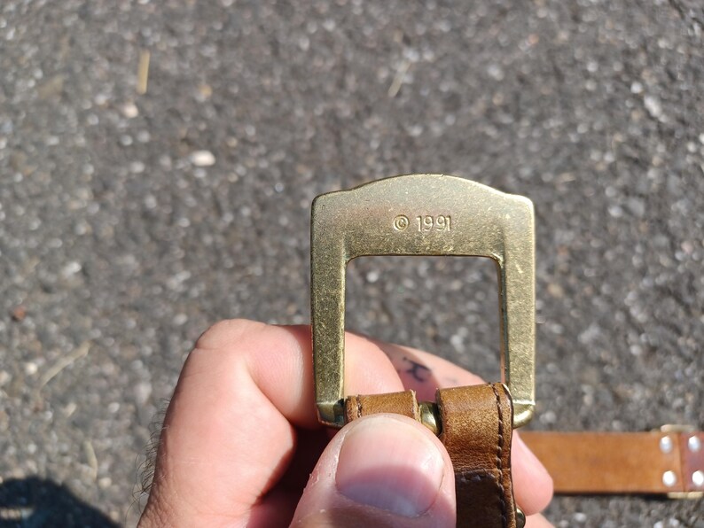 以下が含まれることがあります： 長方形で滑らかな表面を持つ、金色の金属製のベルトバックル。バックルには「&copy; 1991」という小さな刻印があります。
