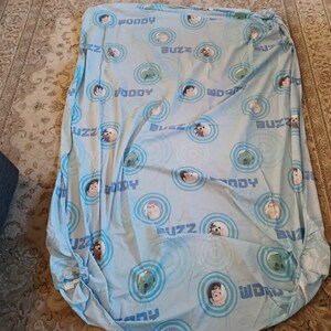 May include: A blue fitted sheet with a repeating pattern of Woody and Buzz Lightyear from the Toy Story movie franchise. The sheet features the characters' faces and the words "Woody" and "Buzz" in a circular design.