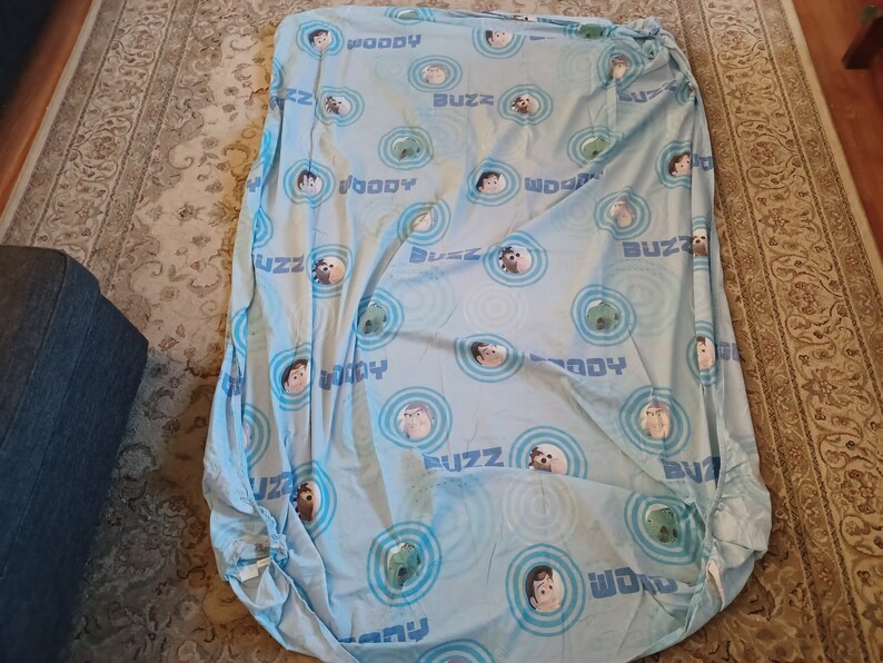 May include: A blue fitted sheet with a repeating pattern of Woody and Buzz Lightyear from the Toy Story movie franchise. The sheet features the characters' faces and the words "Woody" and "Buzz" in a circular design.