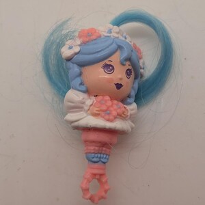 May include: A small plastic figurine featuring a character with light blue hair, a white floral headpiece, and a white dress. The character holds a cluster of pink flowers. The base is a pink, textured, ring-shaped handle with blue accents.