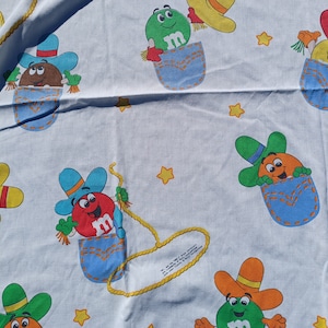 May include: A white fabric with a repeating pattern of colorful M&M's characters dressed as cowboys. The characters are wearing blue jeans, brown cowboy hats, and red bandanas. The background is white with a few scattered stars.