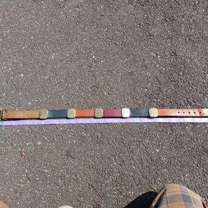 以下が含まれることがあります： 茶色の革製のベルトに真鍮製のバックルと、茶色、黒、赤、金など様々な色の装飾的な金属の正方形が付いている。ベルトは灰色のアスファルトの表面に紫色のメジャーテープと一緒に平らに置かれている。