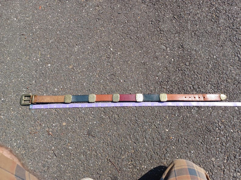 以下が含まれることがあります： 茶色の革製のベルトに真鍮製のバックルと、茶色、黒、赤、金など様々な色の装飾的な金属の正方形が付いている。ベルトは灰色のアスファルトの表面に紫色のメジャーテープと一緒に平らに置かれている。