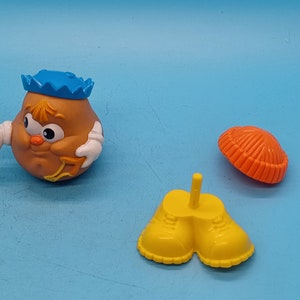 May include: A brown potato head toy with a blue hat, white arms, and a yellow and orange outfit. The toy is part of a set of interchangeable parts for a potato head toy.