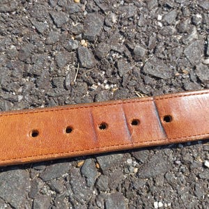 以下が含まれることがあります： 5つの穴が均等に開いた茶色の革製のベルト。ベルトは灰色のアスファルトの表面に置かれています