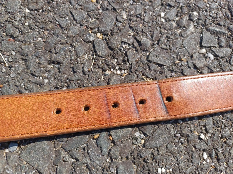 以下が含まれることがあります： 5つの穴が均等に開いた茶色の革製のベルト。ベルトは灰色のアスファルトの表面に置かれています