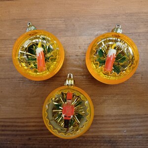 May include: Three vintage Christmas ornaments with a gold glitter background. One ornament features a red and white candle, another features a red and white candle, and the third features a toy soldier.