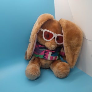 May include: A brown plush bunny wearing white sunglasses with red lenses and a colorful patterned shirt.