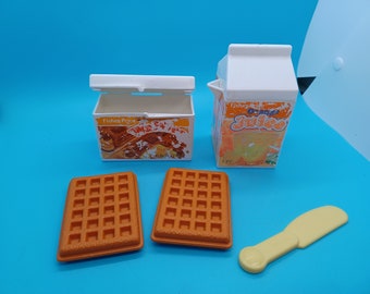 Puede incluir: Un juego de comida de imitación Fisher-Price con dos piezas de waffle naranja, un cuchillo de mantequilla amarillo, un cartón blanco de jugo de naranja que dice "Fisher-Price Jugo de Naranja", y una caja blanca con una imagen de waffles que dice "Fisher-Price Waffles & Syrup".