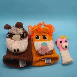 May include: Three knitted hand puppets. The first puppet is brown with white accents and a horse face. The second puppet is orange with white accents and a lion face. The third puppet is pink with white accents and a pig face.