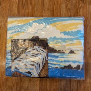 May include: A rectangular blanket sealed in clear plastic packaging. The blanket features a printed landscape scene with a blue ocean, dark rocky coastline, and a yellow and white sky. A product insert card shows the blanket draped over a bed.