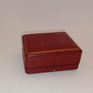 May include: A small, rectangular, red jewelry box with a textured surface. The box has a gold-colored, zig-zag trim around the top edge and a small, gold-colored clasp on the front. The box is closed.