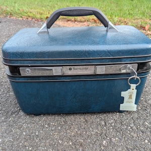 May include: A blue Samsonite hard-shell train case with a textured finish, a black top handle, and a silver-toned metal latch closure. A key is inserted into the lock, and a white tag with the number one is attached to the key ring.