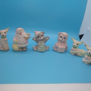 May include: A collection of pastel-colored Christmas figurines, including Santa Claus, reindeer, and teddy bears. The figurines are in various poses, with details like hats, scarves, and a sack. The overall theme is festive and whimsical.
