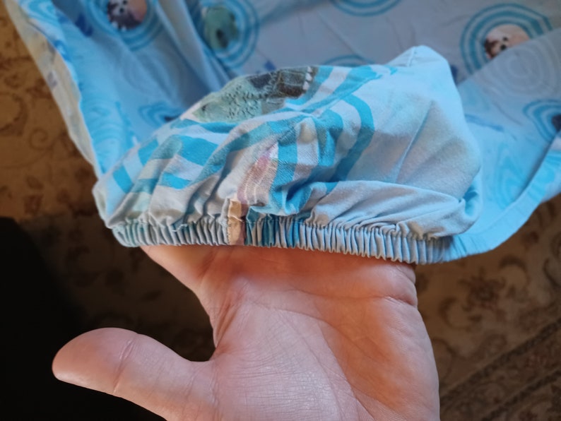 May include: Close-up of a blue and white fitted sheet with a colorful pattern. The sheet has a gathered elastic edge.