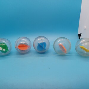 May include: Five clear plastic bouncy balls with different colored shapes inside. The shapes are green, orange, blue, orange, and yellow.