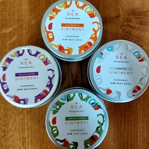 May include: Four small, round, silver tins with colorful designs and labels. Each tin contains a different herbal ointment: Wild Thyme, Calendula, Spruce Resin, and Yarrow. The labels all feature the brand name "REA" and the text "Reviving Folk Wisdom".