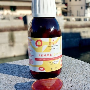Puede incluir: Una botella de vidrio marrón con una tapa blanca y una etiqueta amarilla. La etiqueta dice "REA Reviving folk wisdom HANDMADE FEMME DIVINE THERAPEUTIC MASSAGE OIL 100% NATURAL 100% WILD 100 ml". Hay una piedra roja en forma de corazón en primer plano.