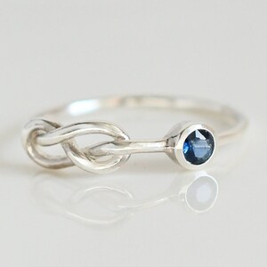 Könnte beinhalten: Ein silberner Ring mit einem Knotendesign und einem kleinen blauen Edelstein. Der Ring liegt auf einem weißen Hintergrund.
